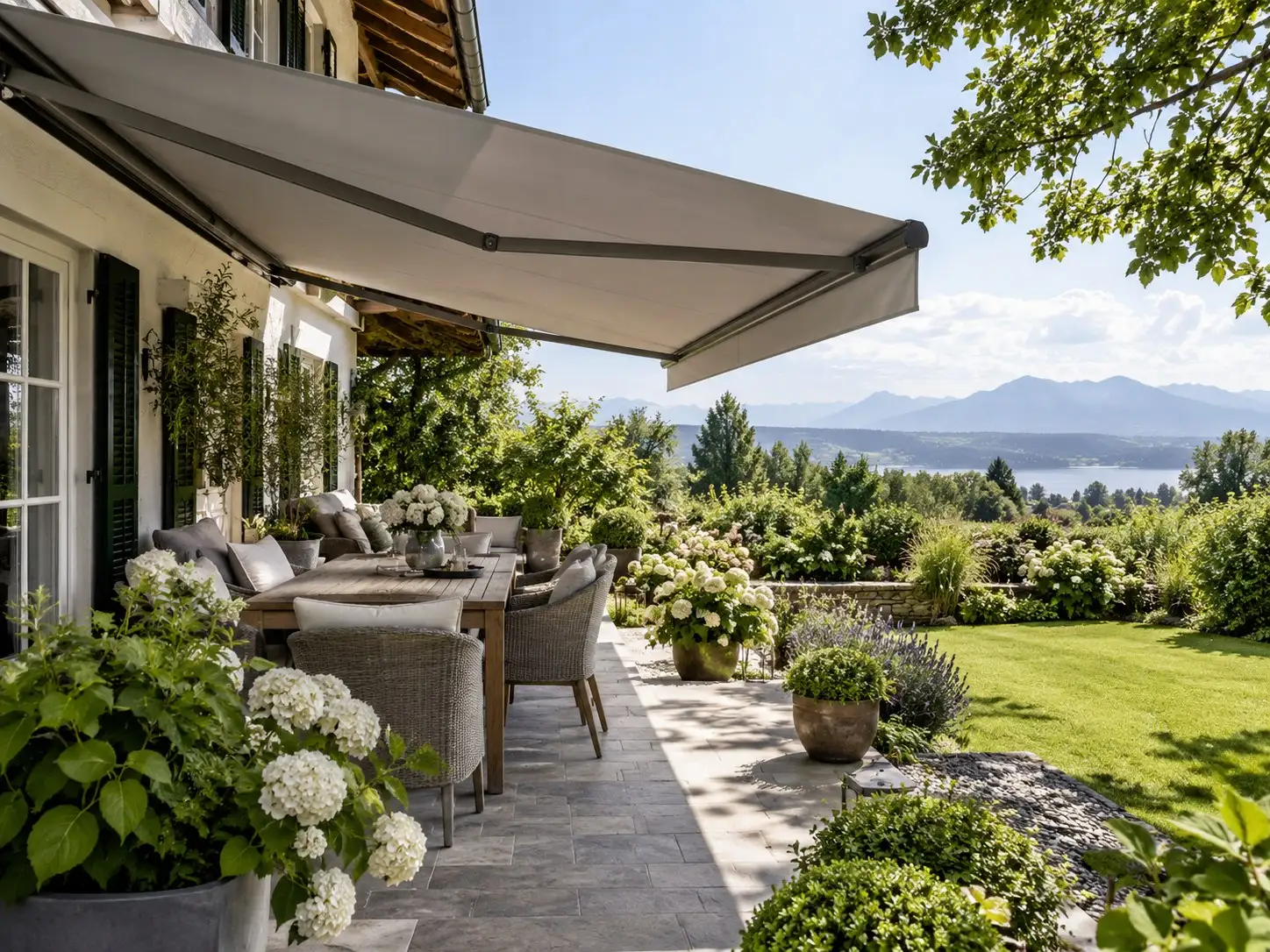 Terrasse mit ausgefahrener grauer Markise, Gartenmöbeln und Hortensien, Blick auf See und Berge